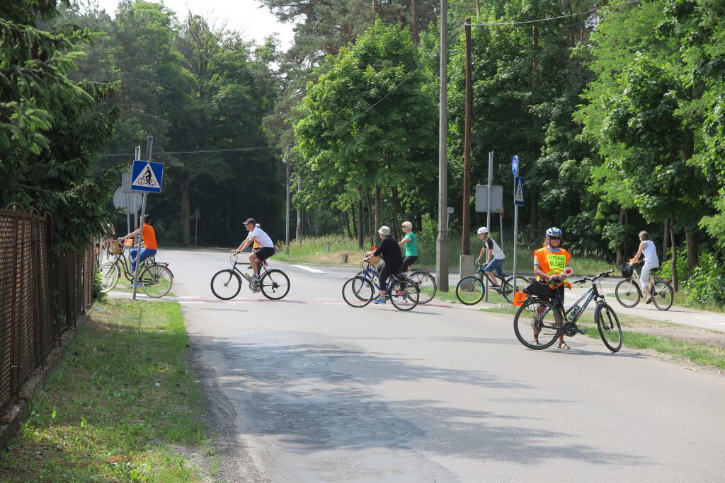 Na pierwszym planie stoi osoba w żółtej kamizelce, za nią inni ludzie przejeżdżają rowerami przez ulicę. 