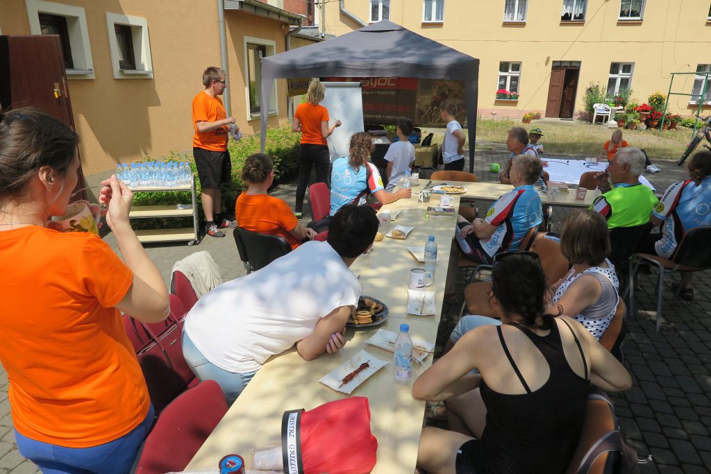 Grupa osób siedzi przy stole. W tle dwie osoby w pomarańczowych koszulkach stoją przy tablicy. 